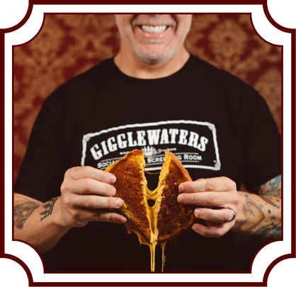 Smiling person wearing a Gigglewaters t-shirt holding a grilled cheese sandwich with melted cheese stretching down, against a decorative burgundy wallpaper background. Image has a vintage-style decorative frame border.