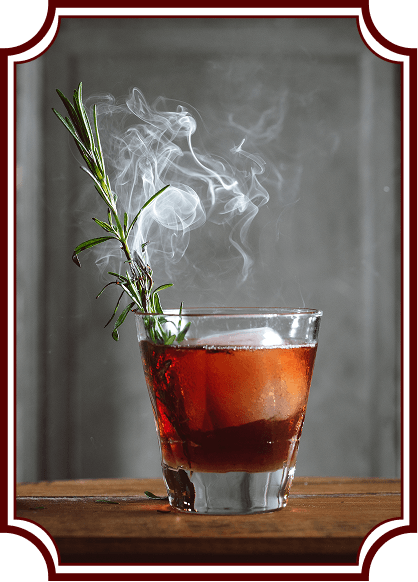 Smoking Rosemary Cocktail A red-amber cocktail in a rocks glass on a wooden surface, garnished with fresh rosemary that's smoking, creating an artistic plume of white smoke rising above the drink. The image has a vintage-style decorative frame border.