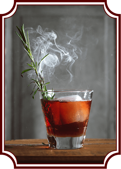 A red-amber cocktail in a rocks glass on a wooden surface, garnished with fresh rosemary that's smoking, creating an artistic plume of white smoke rising above the drink. The image has a vintage-style decorative frame border.