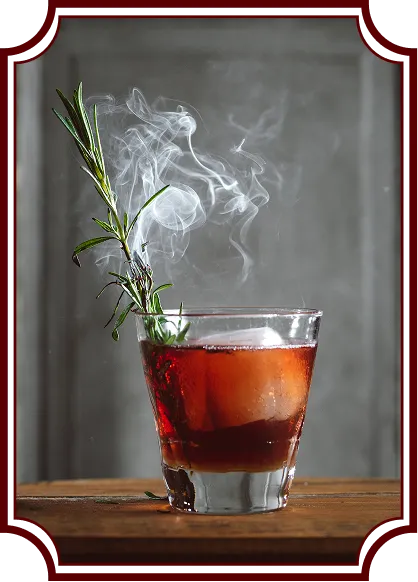 A red-amber cocktail in a rocks glass on a wooden surface, garnished with fresh rosemary that's smoking, creating an artistic plume of white smoke rising above the drink. The image has a vintage-style decorative frame border.