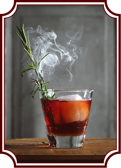 A red-amber cocktail in a rocks glass on a wooden surface, garnished with fresh rosemary that's smoking, creating an artistic plume of white smoke rising above the drink. The image has a vintage-style decorative frame border.